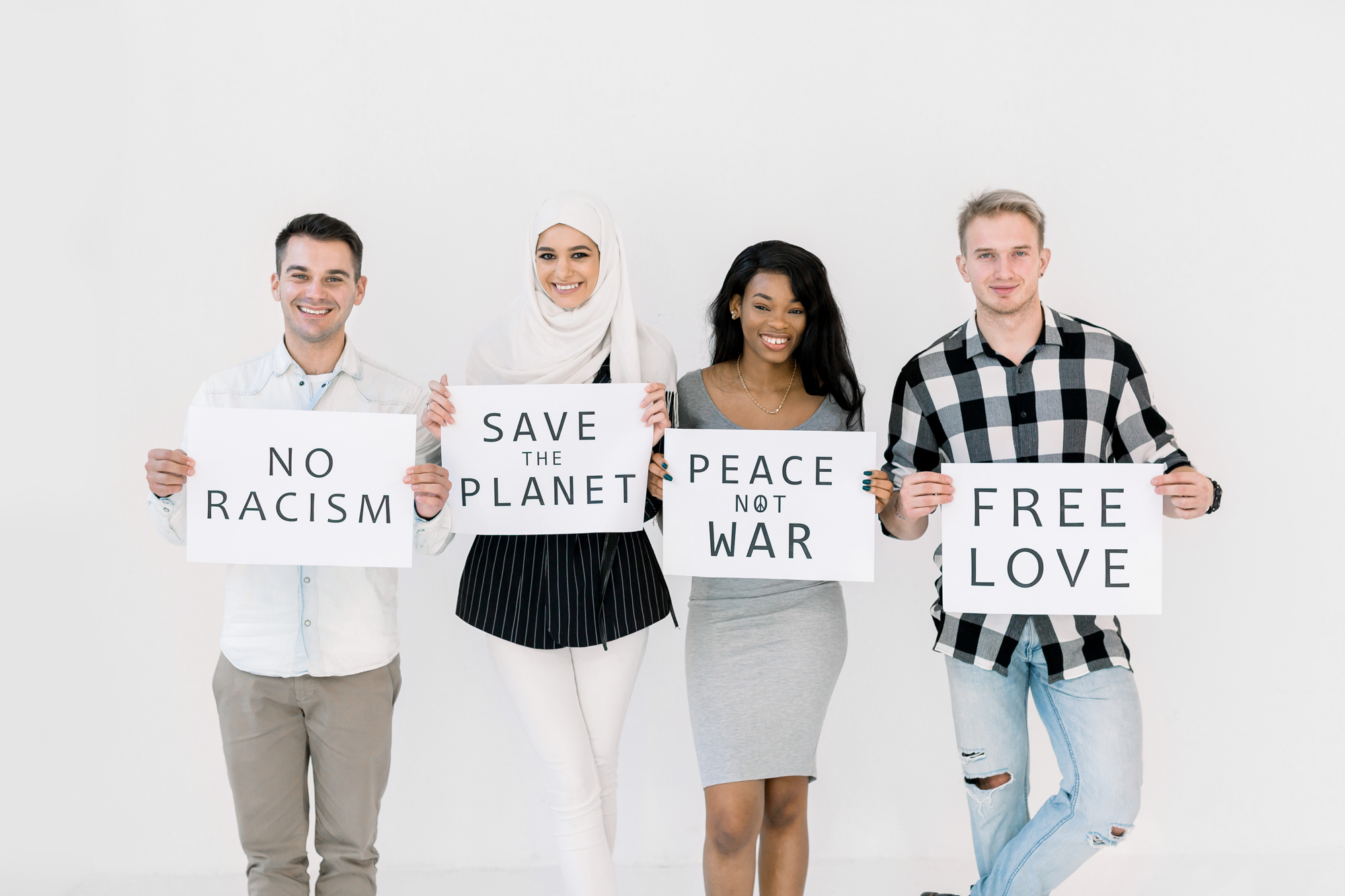 Photo of four cheerful young multiracial friends posing isolated over white background, holding paper sheets with no racism, no war, save the planet text. The concept of peace on earth, equality